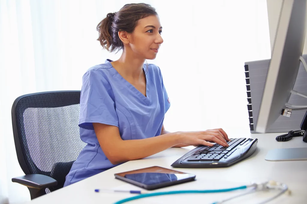 A woman wearing scrubs sits at a desk, typing on a keyboard, and looking at her computer monitor