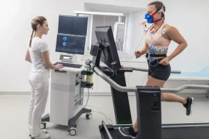 A woman running on a treadmill has medical equipment attached to her as a medical professional monitors their information on a computer monitor